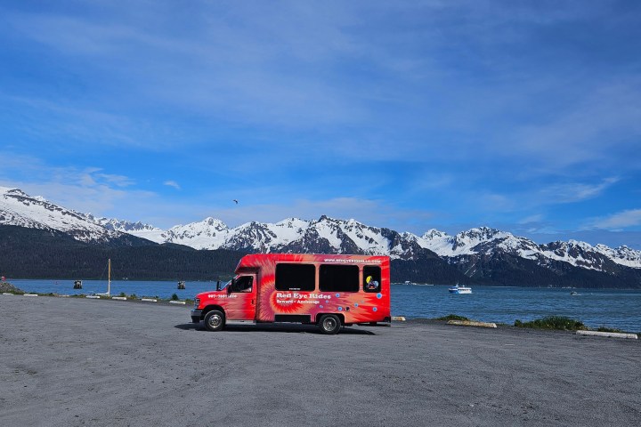 Red shuttle bus by a lake with snowy mountains in the background.
