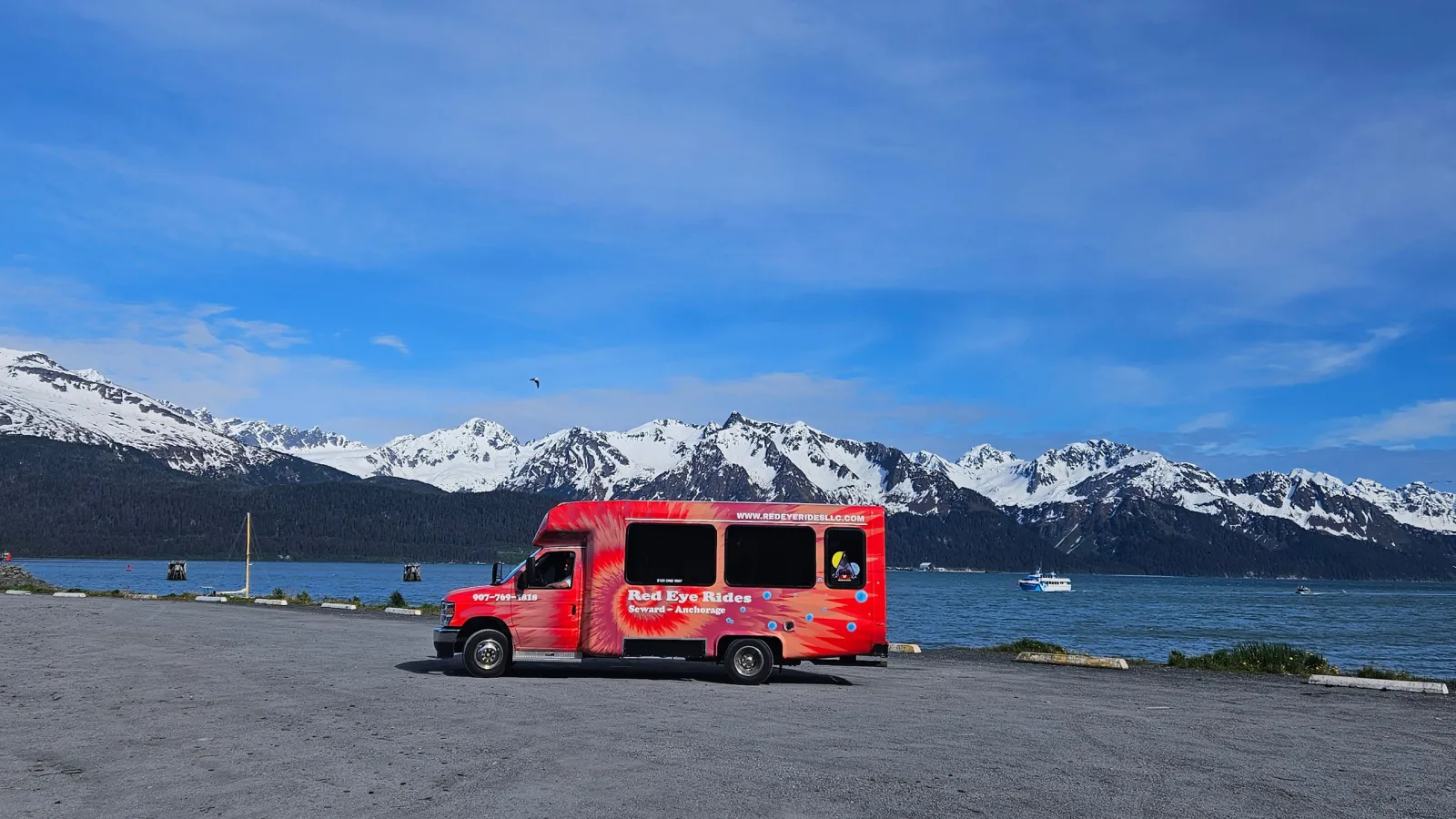 Red shuttle bus by a lake with snowy mountains in the background.