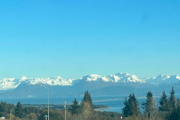 Snow-capped mountains and blue lake under clear blue sky, with trees in the foreground.