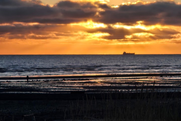 Sunset over ocean with ship on horizon and sandy beach in foreground.