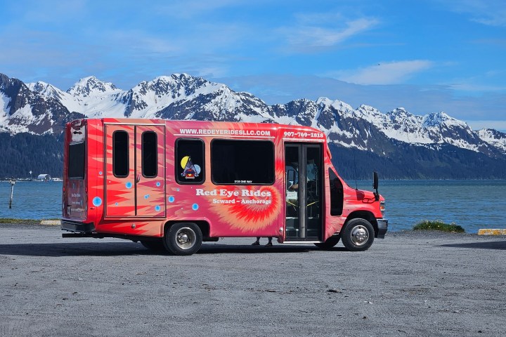 Red van parked by a lake with snowy mountains in the background.