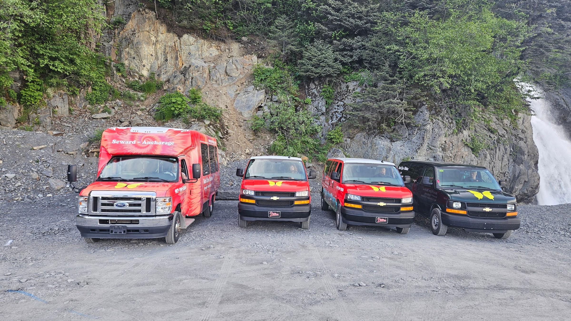 Four colorful vans parked on gravel near rocky cliff and trees.
