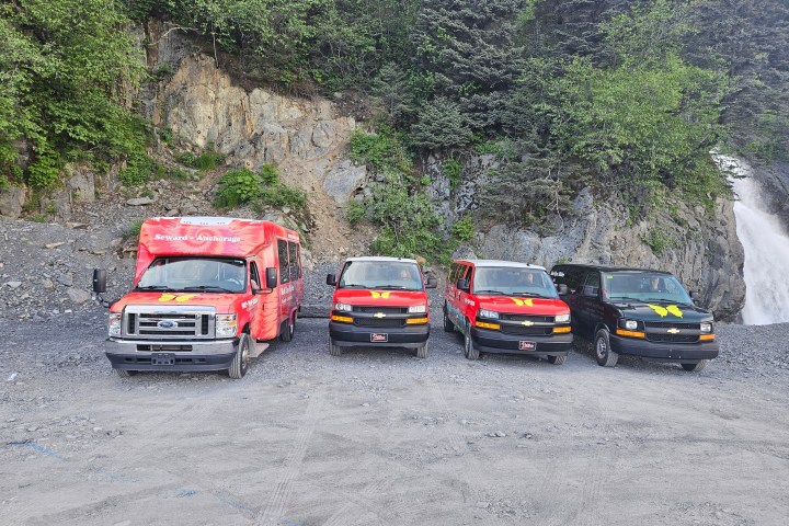 Four colorful vans parked on gravel near rocky cliff and trees.