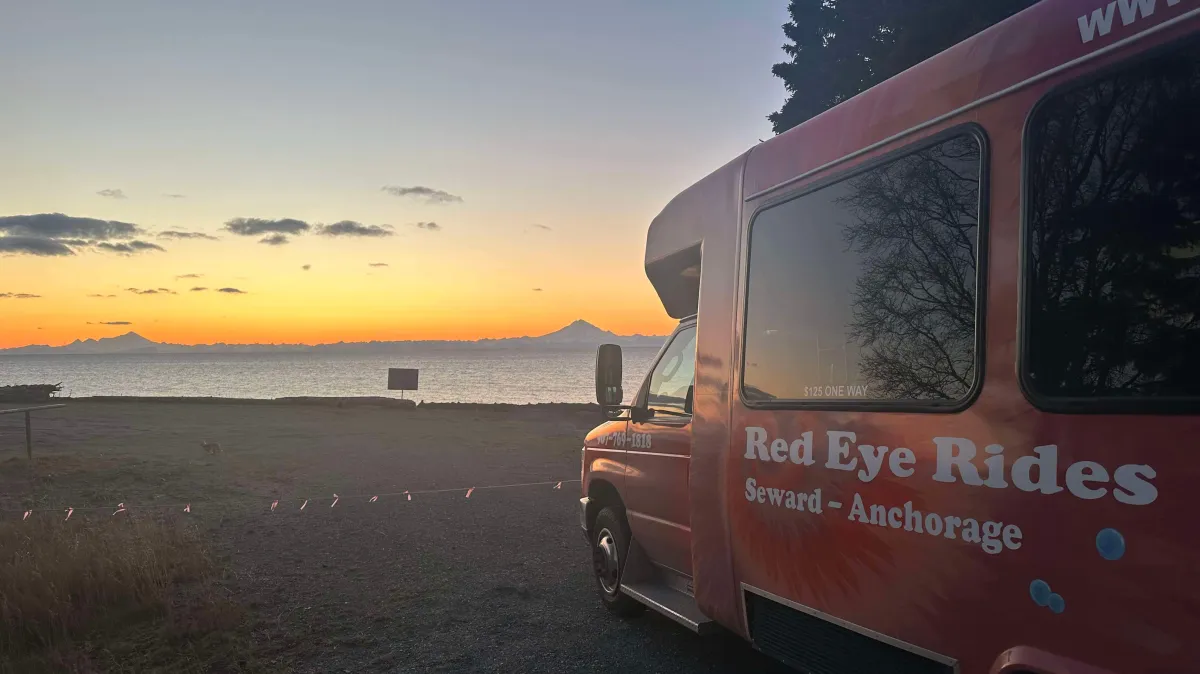 Sunset over calm sea with van, trees, and distant mountains.