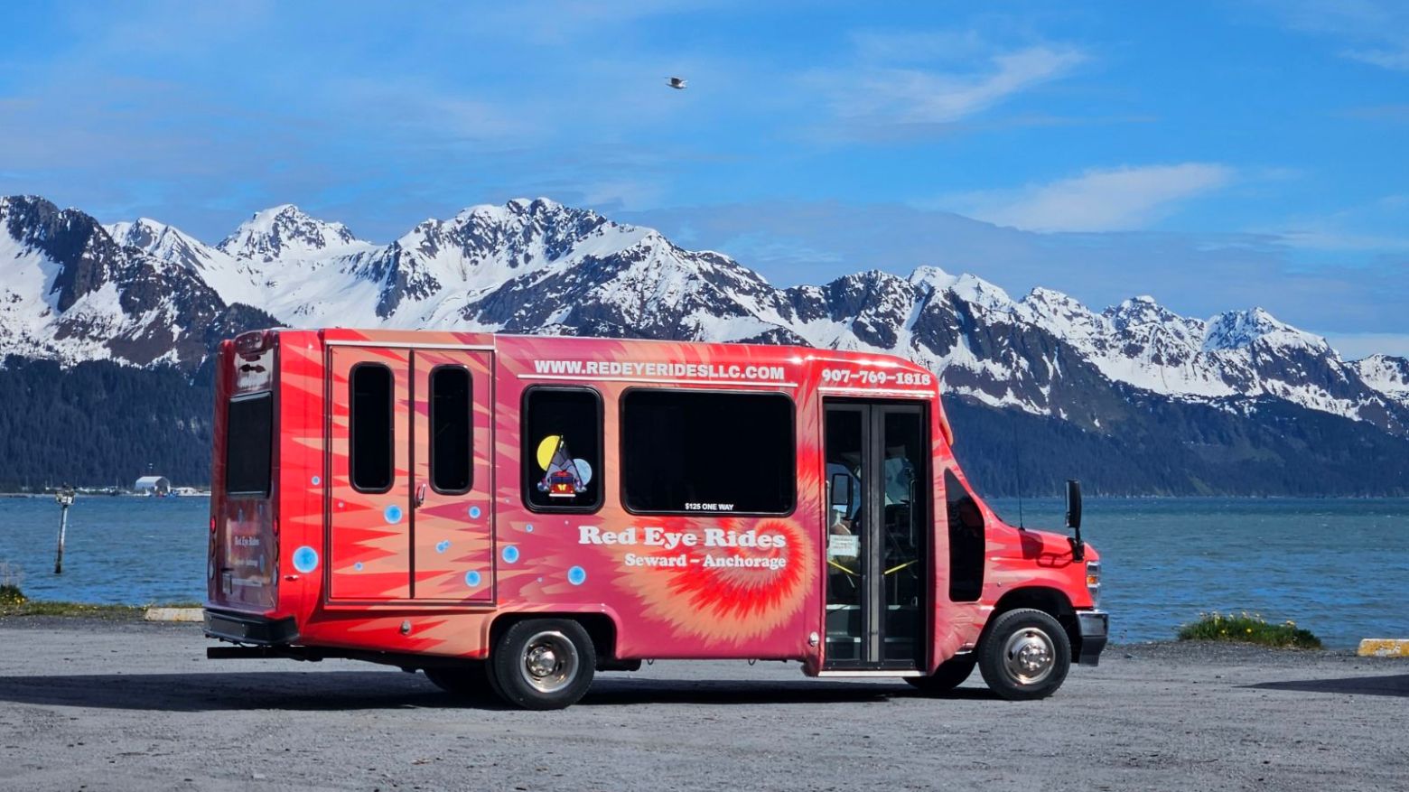 Red shuttle bus parked by a lake and snowy mountains.