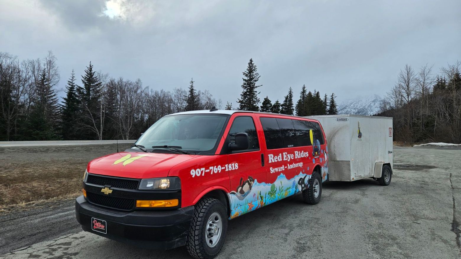 Red van with trailer in mountain area, cloudy sky.